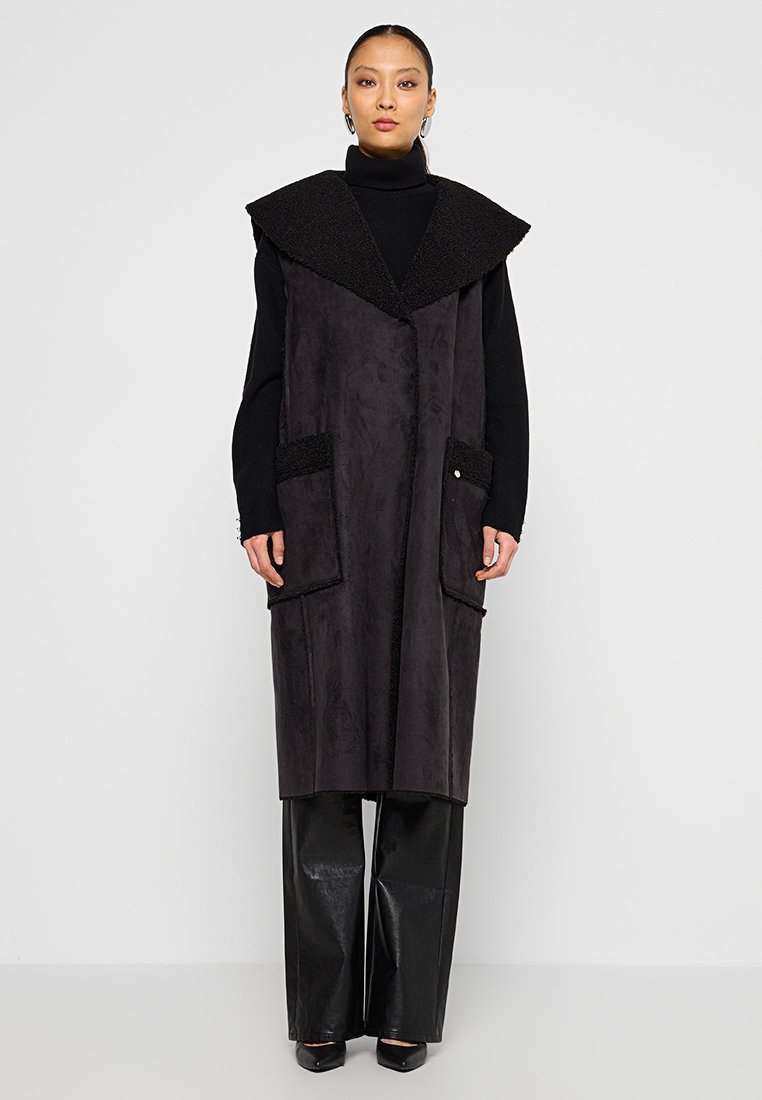 Langer schwarzer Weste mit breitem Kragen, aus weichem Material gefertigt, mit zwei großen Taschen mit strukturierten Akzenten und einer schlanken Silhouette.