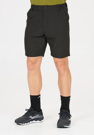 Mann trägt schwarze Sportshorts, schwarze Kniestrümpfe und schwarze Laufschuhe und steht vor weißem Hintergrund.