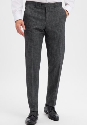 Man wearing dark gray textured dress pants, black leather dress shoes, and a white shirt with one hand in pocket.