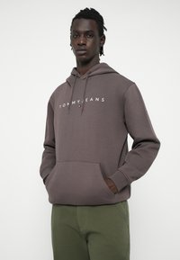 Brūna kapuce ar baltu "TOMMY JEANS" logotipu, regulējamā aukliņā, priekšējā kabata un garās piedurknes, valkāta ar zaļām biksēm.