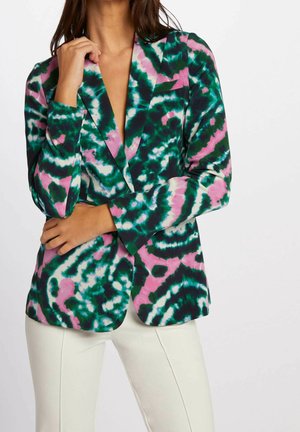 Nainen, joka pukeutunut vihreään, vaaleanpunaiseen ja valkoiseen tummuvaväriseen bleiseriin ja beigenvärisiin housuihin, poseeraa yhdellä kädellä ristissä ja käsi lähellä kasvoja.