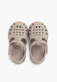 Beige Kinder-Sandalen aus flexiblem Kunststoff, mit einem geflochtenen Design, offenen Seiten und einem Riemen an der Ferse für einen sicheren Sitz.