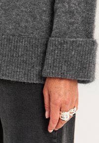 Grijze gebreide trui mouw met geribde manchetten, met een zachte textuur. Hand versierd met drie metalen ringen, vingers tegen een donkere stof.
