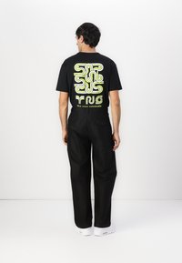 Svart t-skjorte med et komplekst gult-grønt grafisk design på baksiden, kombinert med løse svarte bukser og hvite sneakers.