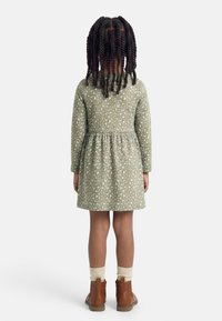 Παιδί με πλεκτά μαλλιά, γυρισμένο με την πλάτη, φοράει μακρυμάνικο φόρεμα με σχέδια, κρεμ κάλτσες και καφέ μποτάκια μέχρι τον αστράγαλο, σε λευκό φόντο.