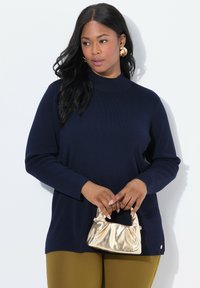 Frau mit langen schwarzen Haaren trägt einen marineblauen Pullover und senfgelbe Hose, hält eine kleine metallisch goldene Handtasche vor einem weißen Hintergrund.