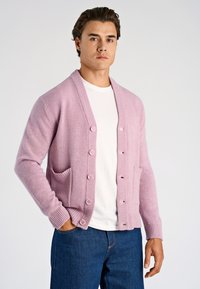 Vaaleanpunainen neuletakki pehmeästä materiaalista, jossa on resorikaulus, kuusi nappia ja kaksi etutaskua. Päällä valkoinen paita ja siniset farkut.