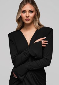 Abito nero a maniche lunghe con scollo a V profondo. Il materiale sembra morbido con un design drappeggiato e maniche a campana. La modella ha le unghie curate e smaltate di bianco.