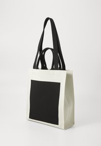 Borsa a spalla in tela di colore crema con un ampio motivo a quadrato nero, doppi manici e cinturini neri a contrasto.