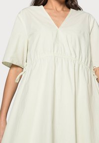 Vaaleanvihreä, lyhythihainen mekko, jossa on V-mallinen kaula-aukko, koottu yksityiskohta vyötäröllä ja solmut sivusaumoissa; valmistettu pehmeästä, virtaavasta kankaasta.