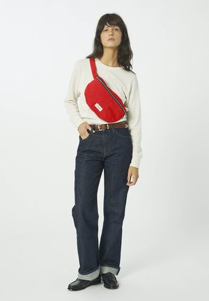 Femme portant une chemise blanche à manches longues, un jean bleu foncé avec des revers, des chaussures noires et un sac bandoulière rouge porté en travers de la poitrine.