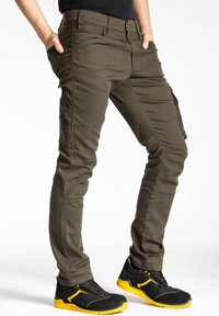 Pantalons cargo vert olive avec une coupe slim, plusieurs poches et un tissu robuste. Associés à des baskets noires avec des accents jaunes.