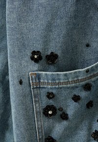 Denimstof med en lys blå nuance, prydet med sorte blomsterdetaljer og palietter, med synlige syninger og en lomme.