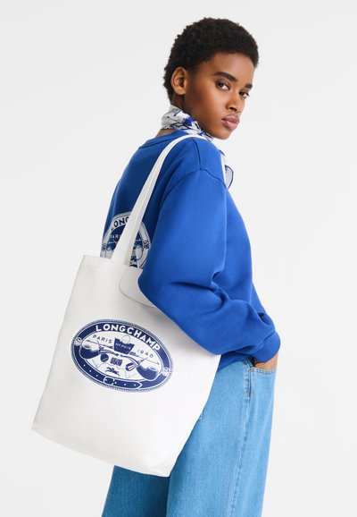 Weiße Canvas-Tragetasche mit blauem Logodesign, ausgestattet mit langen Tragegriffen. Das Model trägt einen blauen Pullover und helle blaue Jeans. Strukturierter Stoff sichtbar.