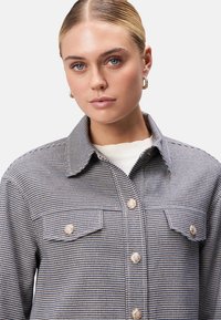 Kariertes Hemd in Blau und Schwarz mit goldenen Beschlägen, zwei Brusttaschen und einem klassischen Kragen; der Stoff hat eine strukturierte Oberfläche.