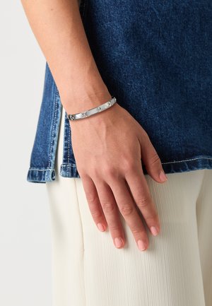 Silberner Metallarmreif mit einem schlanken Design, der geometrische Akzente und eine glänzende Oberfläche aufweist, getragen an einem hellen Hautarm. Denim und plissierter Stoff im Hintergrund.