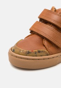 Zapatilla de cuero marrón con dos correas de Velcro ajustables, punta redondeada y una suela de goma texturada con un acento moteado en la puntera.