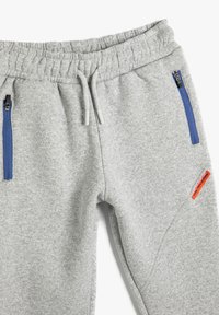Graue Sweatpants aus einem weichen Material, mit einem elastischen Bund, blauen Reißverschlusstaschen und einem orangefarbenen Logopatch an der Seite.