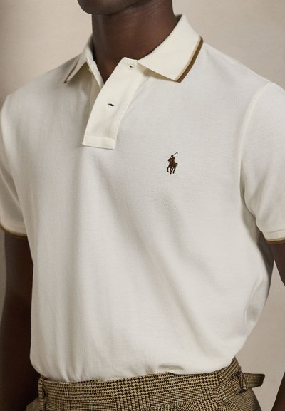 Vyras dėvi kreminės spalvos polo marškinėlį su mažu siuvinėtu polo žaidėjo logotipu ir rudus languotus aukšto juosmens kelnes.