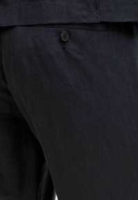 Κοντινό πλάνο σκούρου ναυτικού tailored παντελονιού με πίσω τσέπη με κουμπί και μέρος του τελειώματος ενός ταιριαστού σακακιού ορατό.