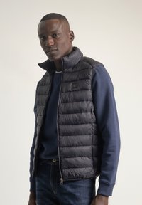 Chaleco acolchado azul marino con canales verticales, cuello alto, cierre con cremallera y un pequeño parche con logo, combinado con un suéter azul marino y jeans azules.