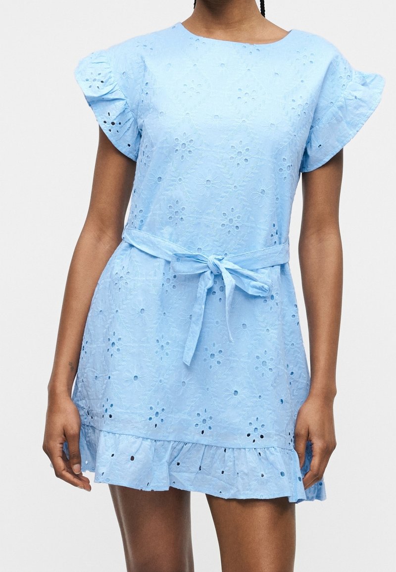 Femme portant une robe bleu clair à œillets avec des manches volantées, une ceinture à la taille et un ourlet, montrée de la nuque à la mi-cuisse sur un fond uni.