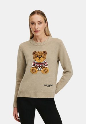 Kvinde iført en beige sweater med en bamse i en mønstret sweater og "Rue Madam Paris" tekst, kombineret med sorte bukser.