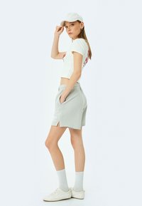 Camiseta blanca recortada con gráfico rojo, pantalones cortos grises con bolsillos, zapatillas blancas y una gorra clara, contra un fondo sólido claro.