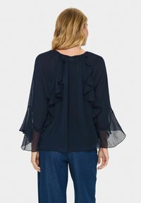 Mörkblå blus med volanger längs ryggen och breda, flödande ärmar. Tillverkad av genomskinligt tyg, ihop med lösa denimbyxor.