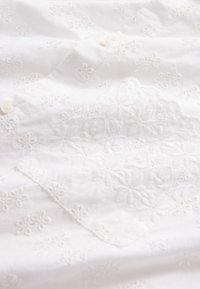 Balta medvilninė medžiaga su siuvinėtais gėlių raštais, puoštais sagų detalėmis ir tekstūruotu paviršiumi. Pakartoti keli gėlių motyvai visoje medžiagoje.