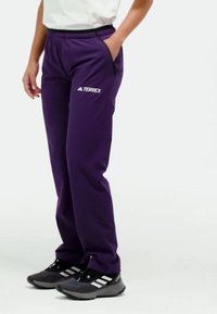 Paarse sportbroek met een elastische tailleband, zijzakken en het "TERREX"-logo in het wit. Gediend met zwart-witte sneakers.