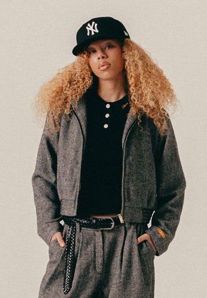 Jeune femme aux cheveux blonds bouclés portant une casquette noire, une chemise noire à boutons et une veste et un pantalon gris à motifs avec une ceinture tressée.