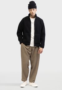 Veste en polaire noire avec fermeture éclair à l'avant, deux poches avant, coupe surdimensionnée, associée à un pantalon texturé beige et des baskets blanches. Bonnet en tricot noir.