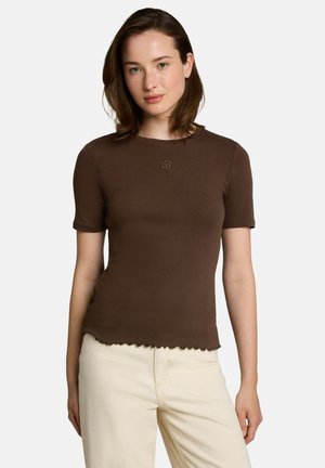 Vrouw met schouderlang bruin haar die een nauwsluitend donkerbruin T-shirt met korte mouwen en lichtbeige broek draagt tegen een neutrale achtergrond.