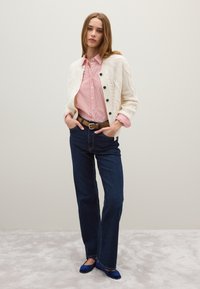 Cremefarbener Strickcardigan mit Zopfmuster und schwarzen Knöpfen, rot-weiß gestreifte Bluse, dunkelblaue ausgestellte Jeans und blaue Ballerinas.