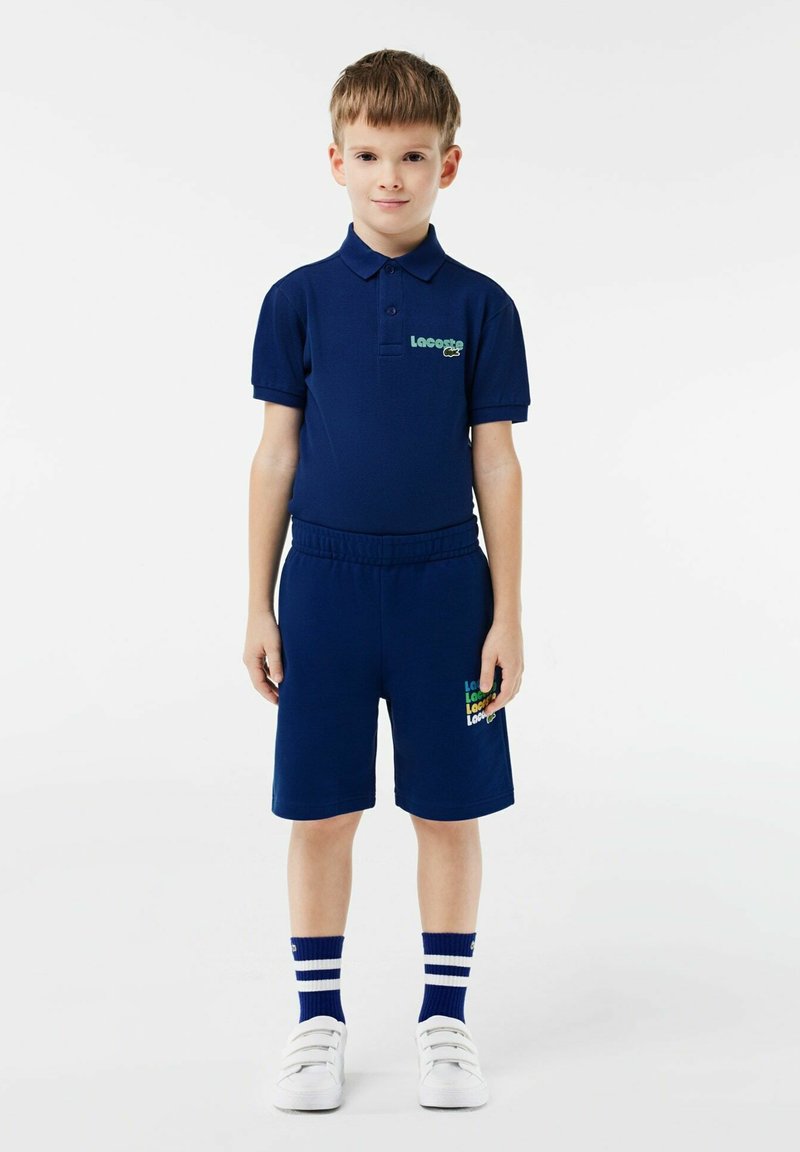 Ragazzo in piedi, vestito con una polo blu scuro e pantaloni corti abbinati, con sneakers bianche e calzettoni blu scuro con righe bianche.