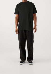 Svart bomullströja med rund hals, kombinerad med löst sittande svarta byxor och svarta sneakers med vita sulor, stående mot en neutral bakgrund.