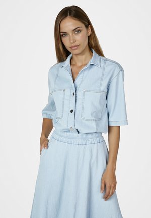 Woman with straight brown hair wearing a light blue short-sleeve denim shirt and matching high-waisted skirt against a white background.