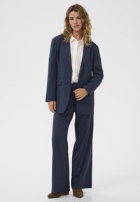 Tengerészkék tűcsíkos öltöny egysoros zakóval és bőszárú nadrággal, fehér inggel és barna velúr bokacsizmával párosítva.