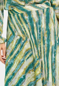 El vestido fluido presenta un patrón de rayas verdes y doradas sobre un fondo claro. La tela parece tener una textura con un suave brillo.
