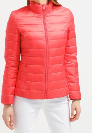 Punainen pehmeä takki korkealla kauluksella, etuvetoketjulla ja ommelluilla vaakasuorilla paneeleilla. Materiaali näyttää sileältä ja kiiltävältä, yhdistettynä valkoisiin housuihin.