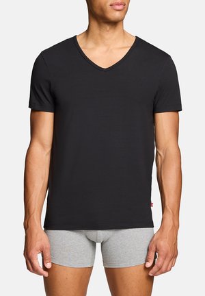 Mann trägt ein eng anliegendes schwarzes V-Ausschnitt-T-Shirt mit kurzen Ärmeln und hellgraue Boxershorts, steht vor einem einfarbigen Hintergrund.