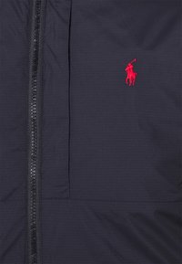 Marinblå vindjacka med en kontrasterande svart dragkedja och en röd polo-logga på bröstet. Har en slät textur och en strömlinjeformad design.