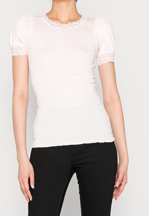 Jasnoróżowa dopasowana bluzka z krótkimi bufkami, wykończona koronką przy dekolcie i rękawach, zestawiona z czarnymi dopasowanymi spodniami. Gładka tekstura.