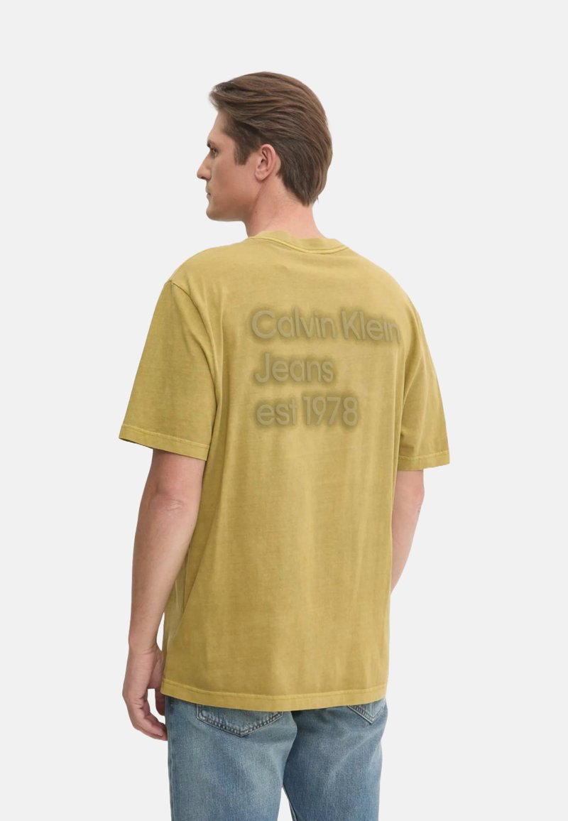 Camiseta oversized de color mostaza con mangas cortas, que presenta un logo ligeramente elevado en la parte posterior: "Calvin Klein Jeans est 1978."