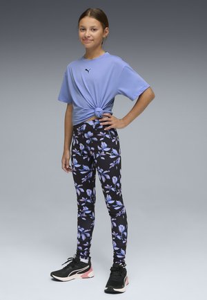 Niña joven de pie con la mano en la cadera, vistiendo una camiseta lavanda de Puma anudada, leggings florales y zapatillas negras sobre un fondo gris.