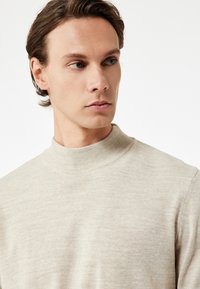 Beige turtleneck neulepaita, jossa on ribattu kaulus, valmistettu pehmeästä ja teksturoidusta kankaasta. Istuva malli, jossa on pitkät hihat.