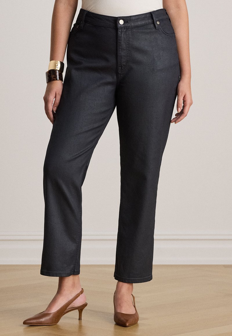 Γυναίκα με μαύρο τζιν με ίσια μπατζάκια, λευκή μπλούζα, καφέ γόβες με λουράκι πίσω και χοντρά βραχιόλια, όρθια σε ξύλινο πάτωμα.