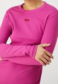 Prodloužený těsný top s dlouhými rukávy v jasně růžovém pruhovaném materiálu, s malým červeným logem "HUGO" na hrudi a kulatým výstřihem.