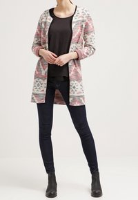 Lang cardigan med et geometrisk mønster i pink, creme og grå over en mørk top og slim fit jeans. Sorte ankelstøvler fuldender outfittet.